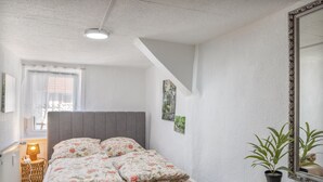Un dormitorio con cama, mesita de noche, lámpara, ventana con cortinas, espejo instalado en la pared y una planta en maceta.