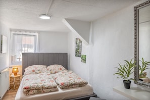 A bedroom with a bed, a bedside table, a lamp, a window with curtains, a wall-mounted mirror, and a potted plant. - Apartment Fewo50 – bis 7 Personen, 2 Schlafzimmer/2 Bäder ab 4 Personen (Homberg (Efze))