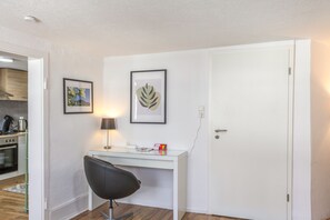 A compact room with a white desk, a chair, a framed leaf artwork, and a door.