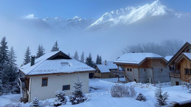 Ein verschneites Bergdorf mit Holzhäusern und freier Sicht auf die schneebedeckten Gipfel im Hintergrund.