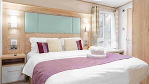 A hotel room with a large bed, wooden headboard, bedside tables, and a window with curtains. - Bella's Retreat - Kelling Heath Holiday Park - 19669 (Holt)