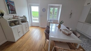 Et moderne køkken med et hvidt bord, stole og trægulv. Der er et vindue med udsigt til grøn vegetation udenfor.