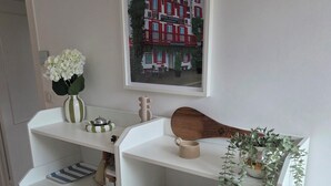 Et hvidt, niveaudelt opbevaringsmøbel med hylder, en hængende plante og et indrammet billede af en bygning.