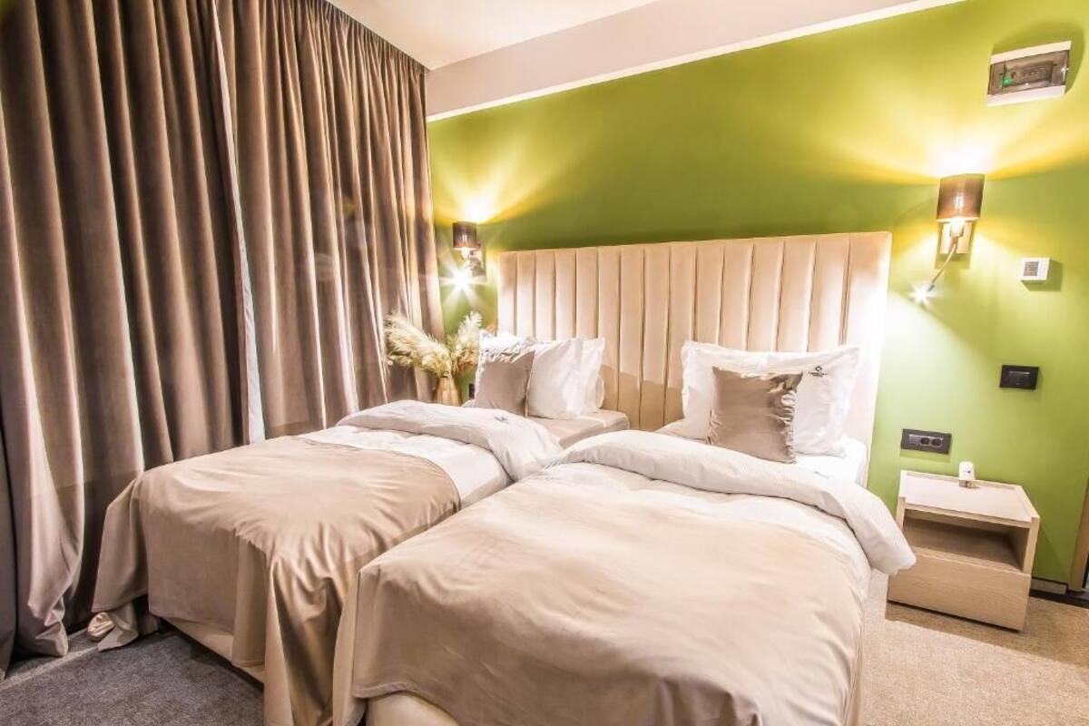 Habitación de hotel con dos camas, una pared de acento verde y una mesita de noche con lámpara.