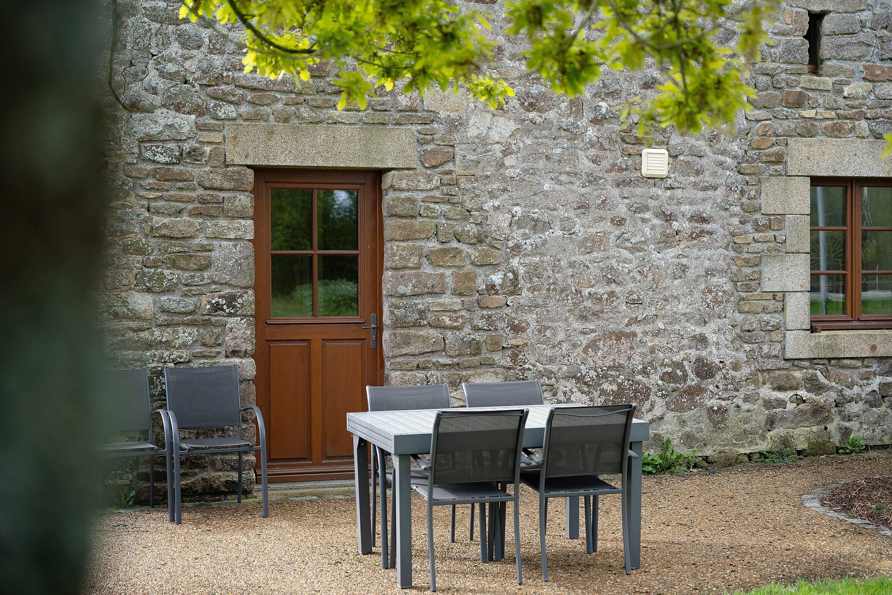 A stone building with a wooden door and windows, a table with chairs, and a gravel ground.