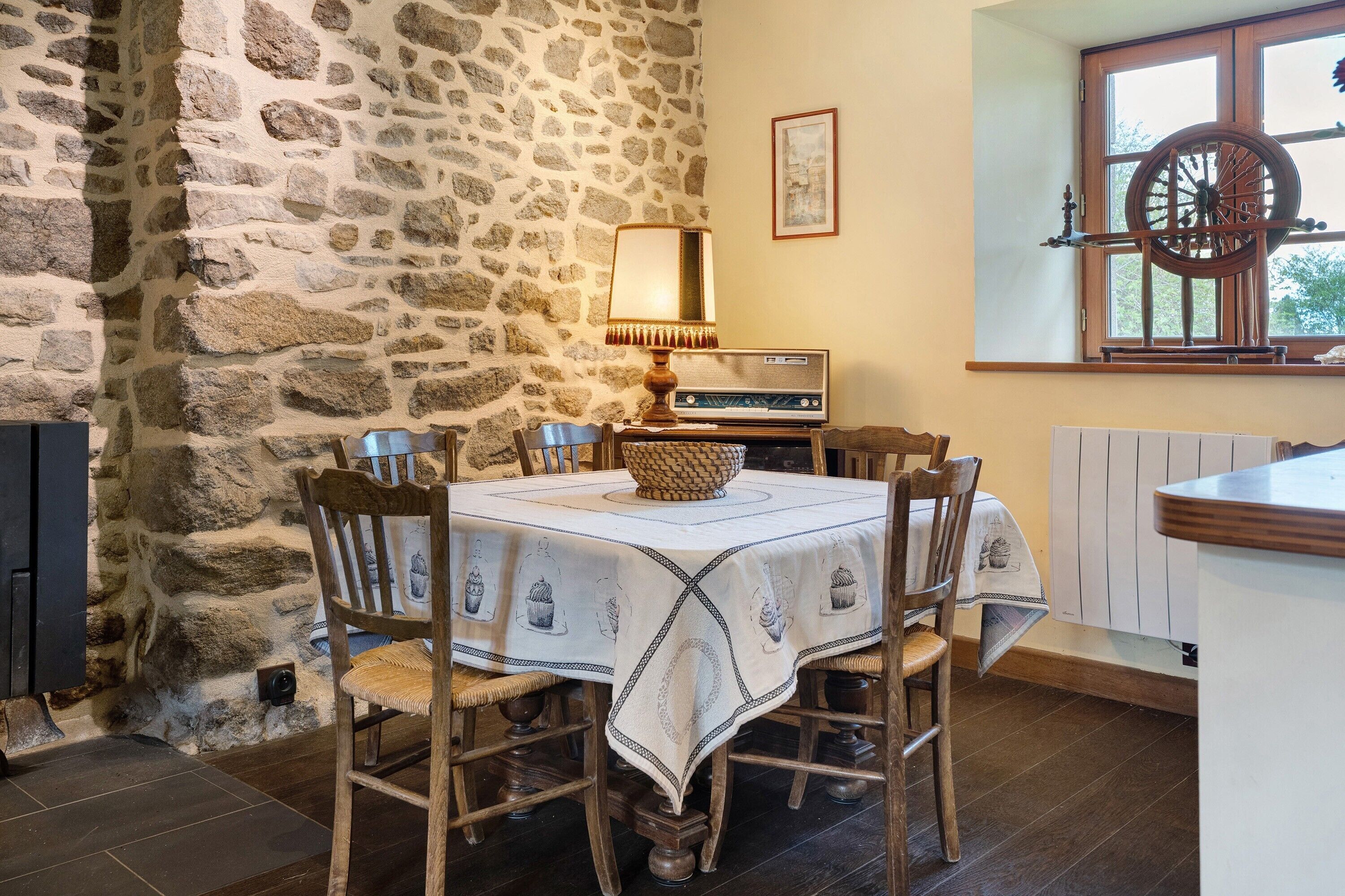 Un mur de pierres, une table recouverte d’une nappe, des chaises, une radio et une fenêtre donnant sur un paysage.