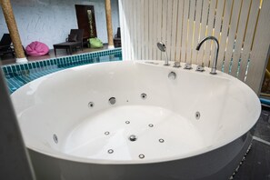 A large, round, white bathtub with multiple faucets and a showerhead, situated in a room with a tiled floor and a view of a pool area with lounge chairs and a door. - Wonderland Amethyst Luxury Villa (Muang Pattaya)