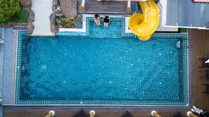 An aerial view of a rectangular swimming pool with a yellow slide, surrounded by a tiled deck and a stone border. - Wonderland Amethyst Luxury Villa (Muang Pattaya)
