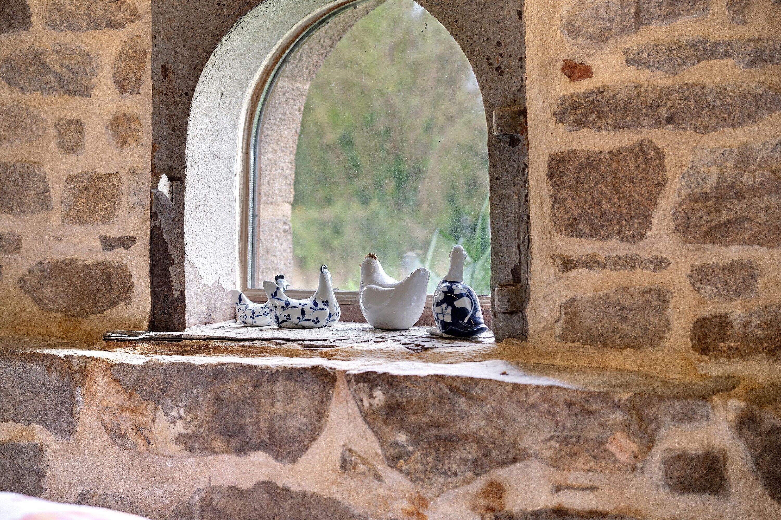 Un mur de pierre avec une fenêtre, une petite étagère ornée d’objets en céramique et une vue sur la verdure à l’extérieur.
