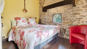 Quarto com cama, cadeira vermelha, parede de pedra e janela com vista.
