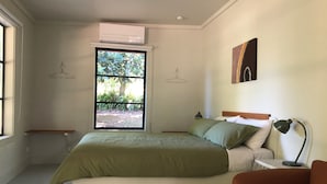 Ein Schlafzimmer mit einem Bett, einem Nachttisch, einer Lampe, einem Fenster mit Blick ins Grüne und einer Wand-Klimaanlage.