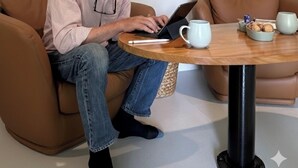 Ein Mann sitzt auf einem braunen Ledersofa und benutzt einen Laptop. Ein runder Holztisch steht daneben, mit einer weißen Tasse und einer kleinen Schale Kekse. Hinter ihm hängen zwei gerahmte Kunstwerke an der Wand.