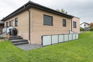 Une maison moderne à deux étages, avec une façade en briques, un porche couvert doté d’une chaise et un petit jardin.