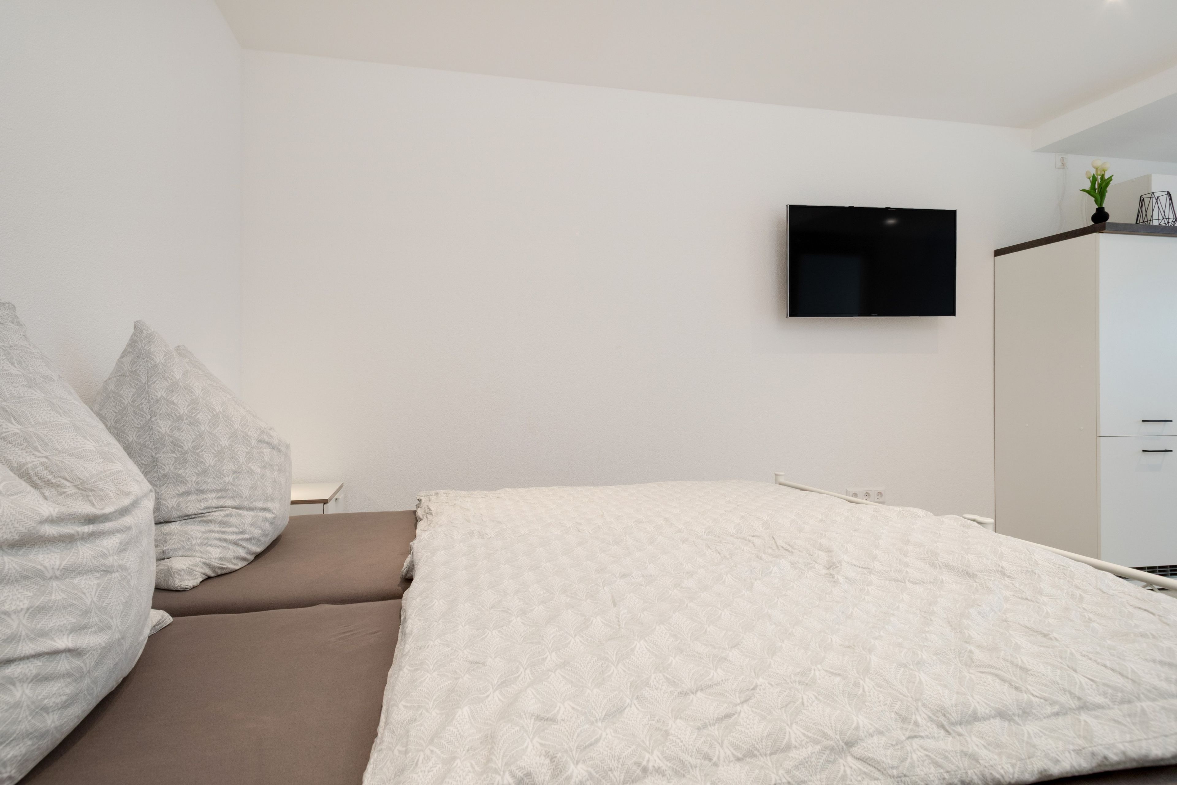 Ein modernes Schlafzimmer mit Bett, Kissen, Fernseher und Kleiderschrank.