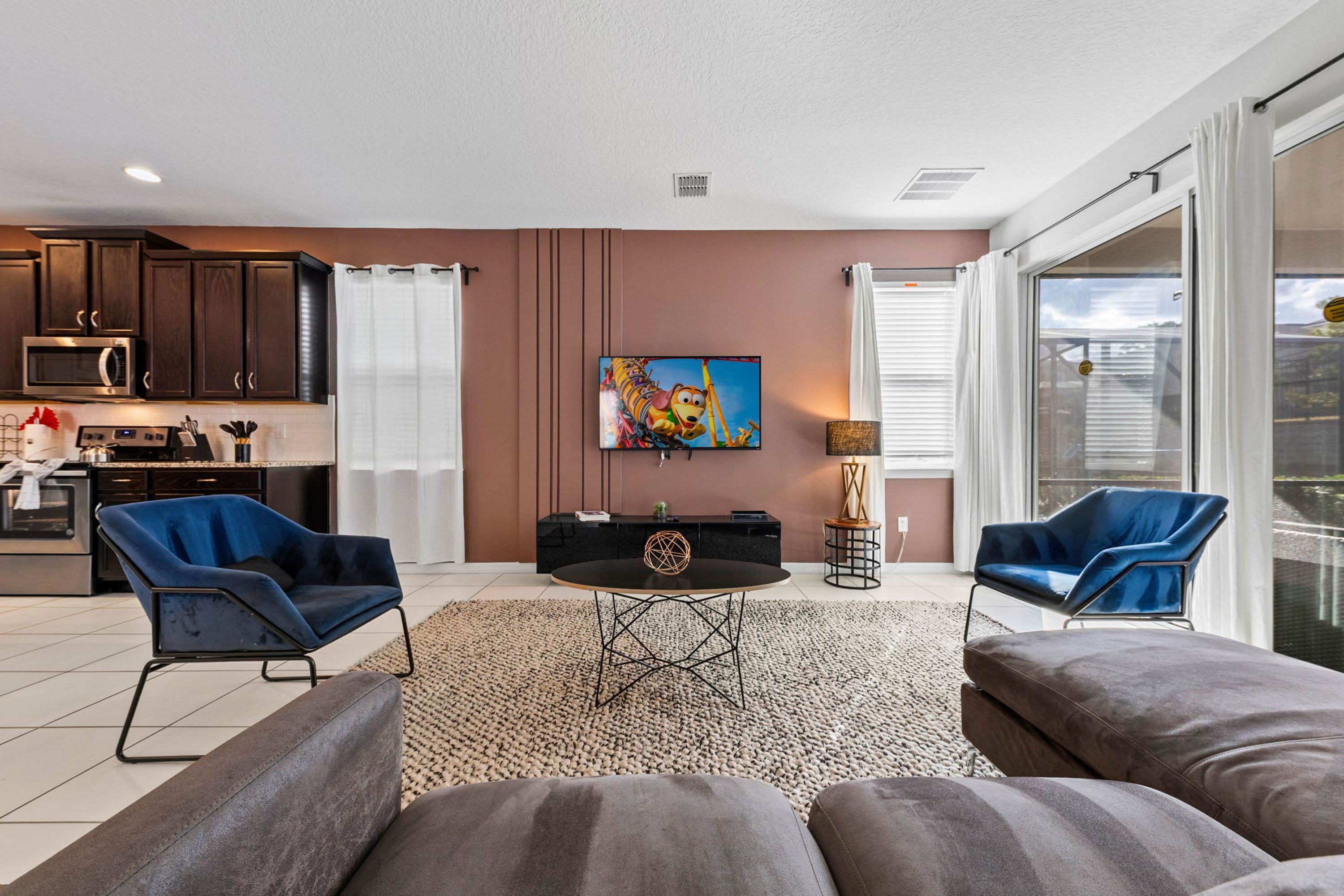 A modern living room with a dining table, two blue armchairs, a sofa, a kitchen in the background, and a large window with curtains.