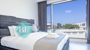 Ein modernes Hotelzimmer mit einem großen Bett, grauem Kopfteil und Blick auf die Stadt durch große Fenster.