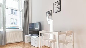 Moderni huone, jossa on suuri ikkuna, litteänäyttöinen televisio, valkoinen työpöytä, kaksi valkoista tuolia ja kaksi kehystettyä kuvaa seinällä.