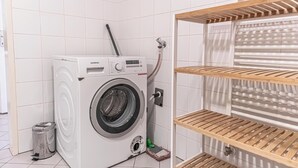 Pesutila, jossa on valkoinen Siemens-pesukone, metallinen roskakori ja puiset hyllyt.