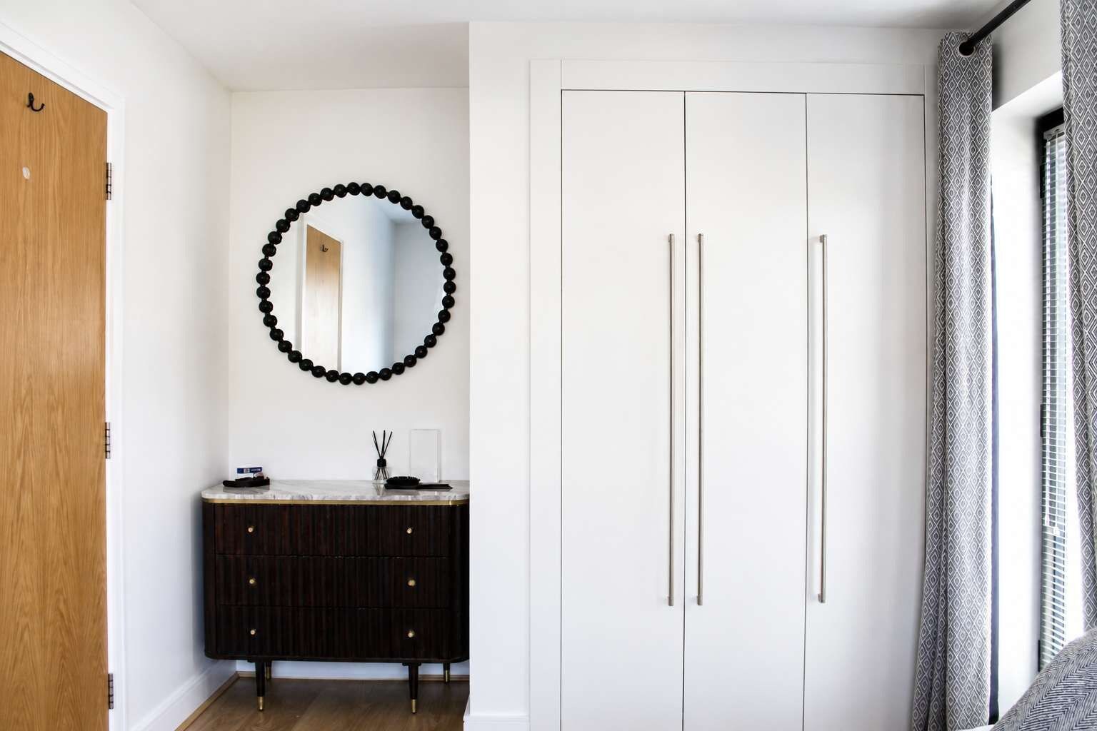Una habitación con puerta de madera, tocador blanco y negro, espejo adornado con una guirnalda de cuentas negras y armario blanco de dos puertas.