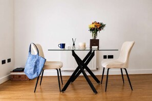 Ein Essbereich mit einem Glastisch, zwei Stühlen, einer blauen Tasche, einer Vase mit Blumen, einer Kerze und einem Buch auf dem Tisch.