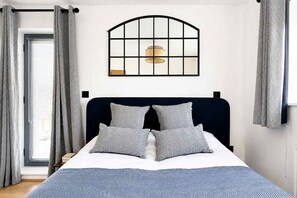 Ein Bett mit dunklem Kopfteil, zwei Kissen, einer blau-weißen Tagesdecke und einem Fenster mit Blick ins Freie.