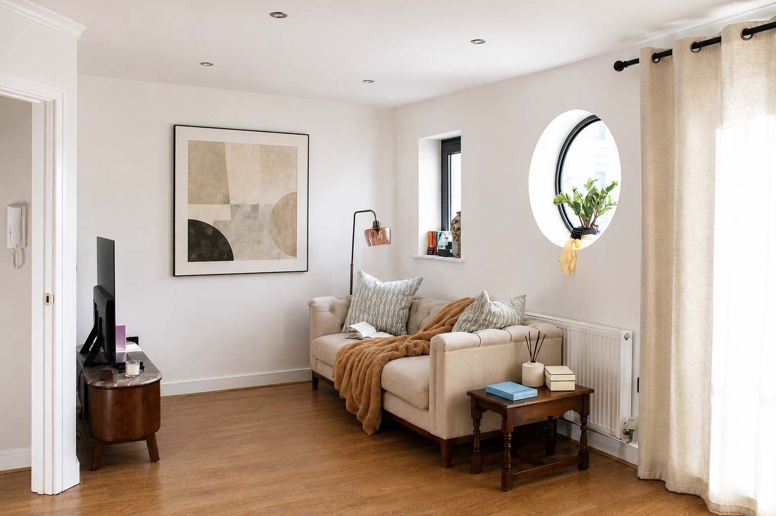 A living room with a sofa, a coffee table, a framed abstract painting, a floor lamp, a round window with a plant, and a radiator.