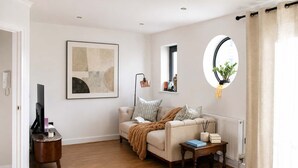 Un salon comprenant un canapé, une table basse, un tableau abstrait encadré, une lampe sur pied, une fenêtre ronde ornée d’une plante et un radiateur.