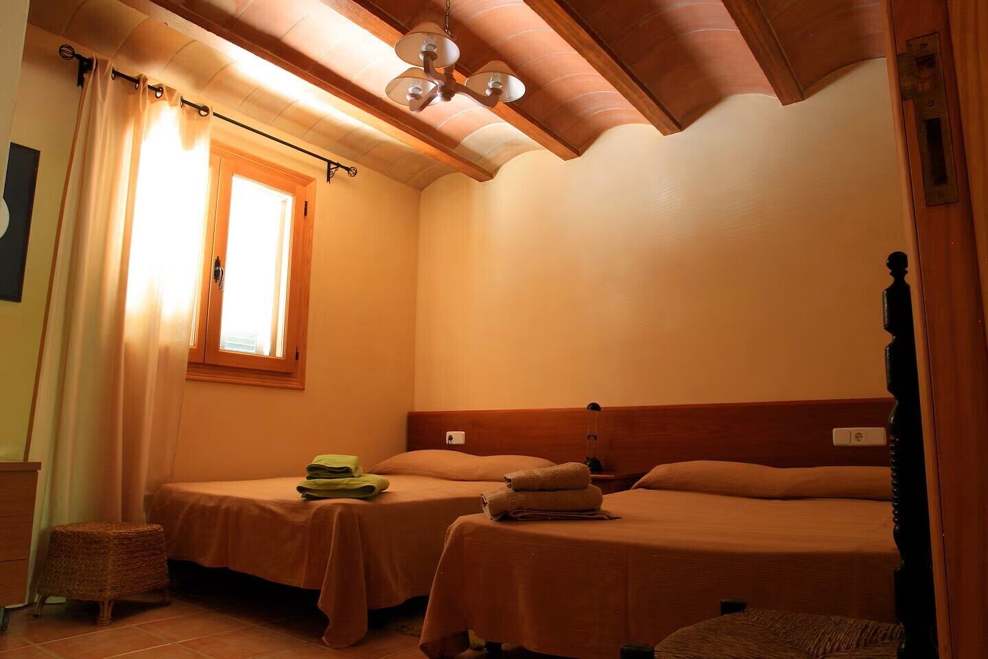 Une petite chambre agréable, dotée de deux lits simples, d’une fenêtre avec des rideaux, d’un plafond en bois et d’une petite table avec une lampe.