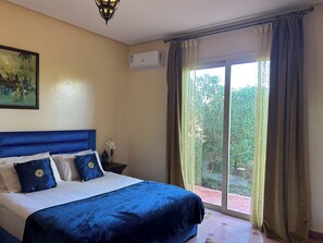 A bedroom with a large bed, a nightstand, a wall-mounted air conditioner, and a window with curtains.