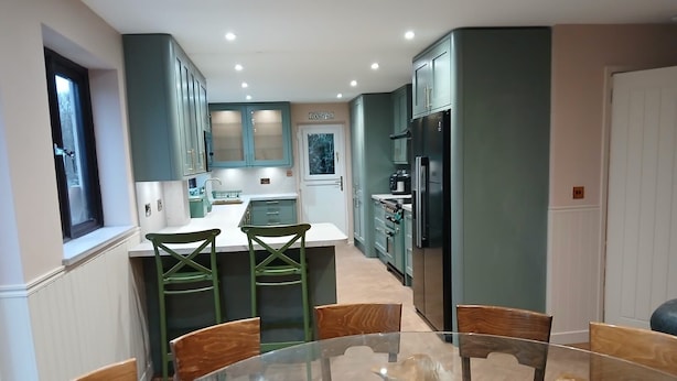 A modern kitchen with teal cabinets, a central island, and a glass dining table surrounded by wooden chairs. - Croydon Lodge (Minehead)