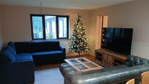 A living room with a blue sofa, a leather couch, a glass coffee table, a wooden TV stand, and a Christmas tree. - Croydon Lodge (Minehead)