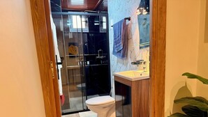 Un baño moderno con cabina de ducha de vidrio, inodoro blanco, tocador de madera con lavamanos y una planta verde grande.