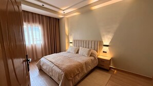 Un dormitorio con una cama grande, una mesita de noche, una lámpara de pared y un ventanal con cortinas.