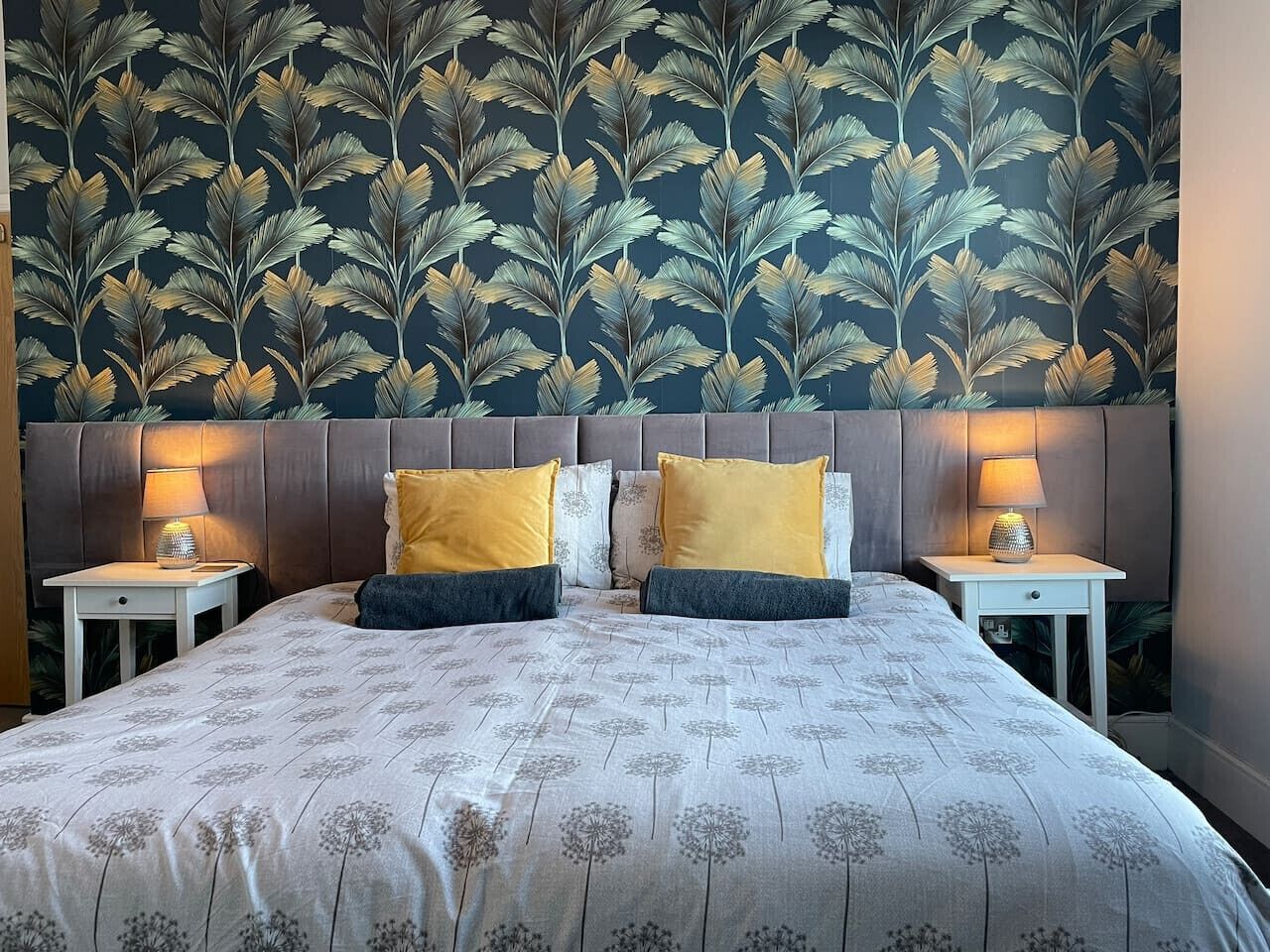 A double bed with a floral bedspread, two bedside tables with lamps, and a dark wallpaper with a leaf pattern.