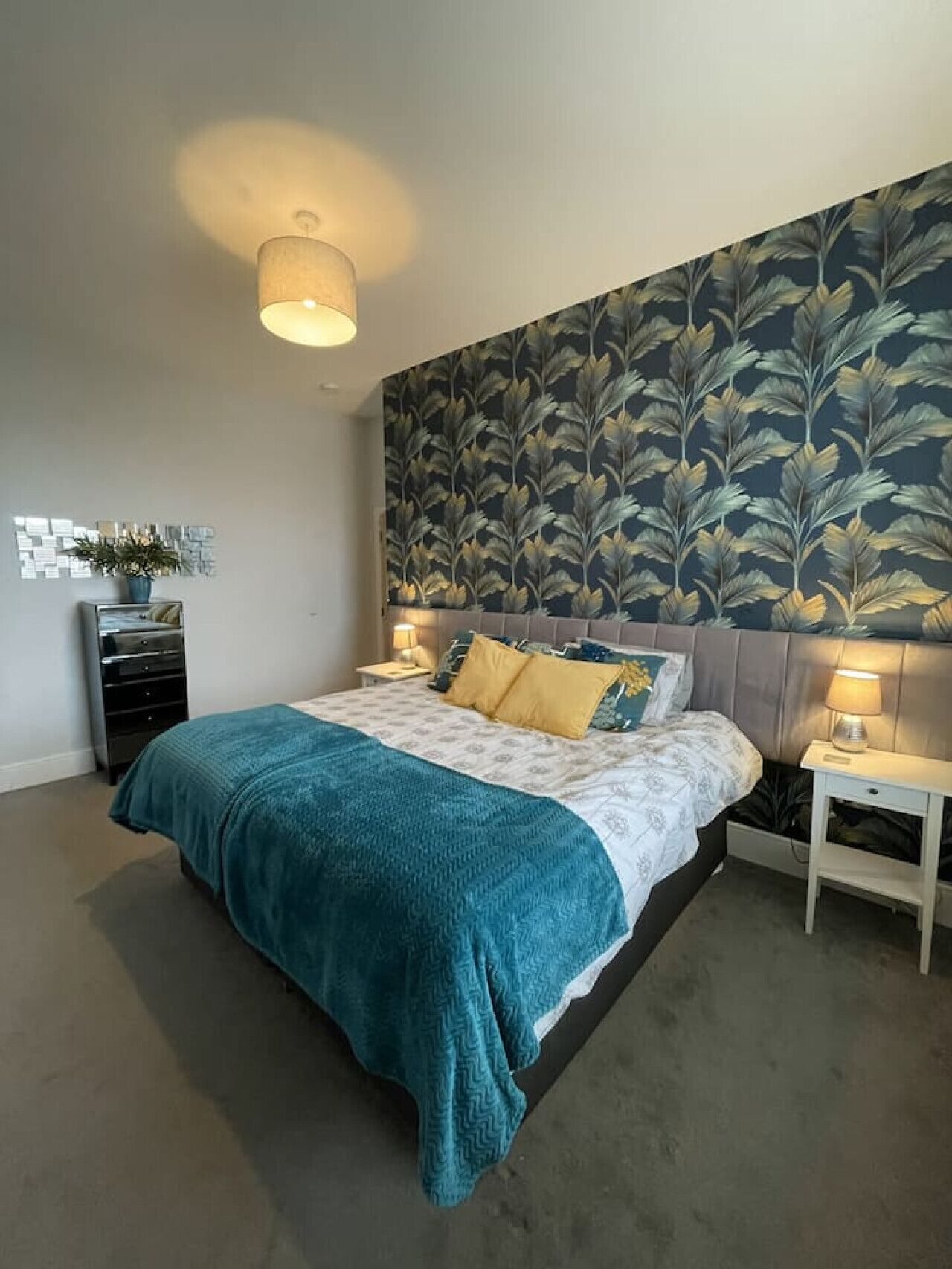 Een slaapkamer met een bed, nachtkastje, kaptafel en een muur met een bladpatroon.