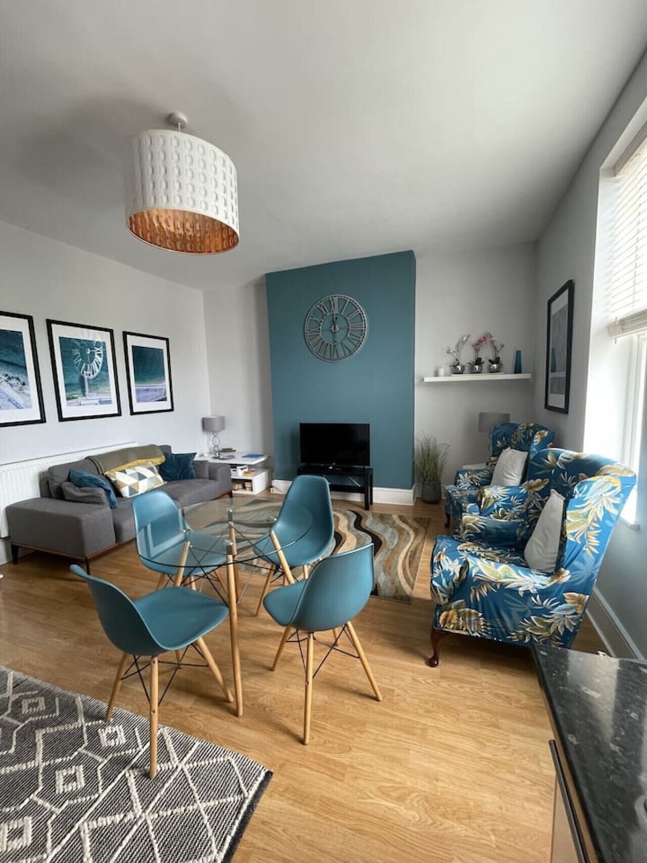 A modern living room with a blue accent wall, a round clock, a flat-screen TV, a sofa, two blue chairs, and a patterned rug.