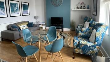 A modern living room with a blue accent wall, a round clock, a flat-screen TV, a sofa, two blue chairs, and a patterned rug.