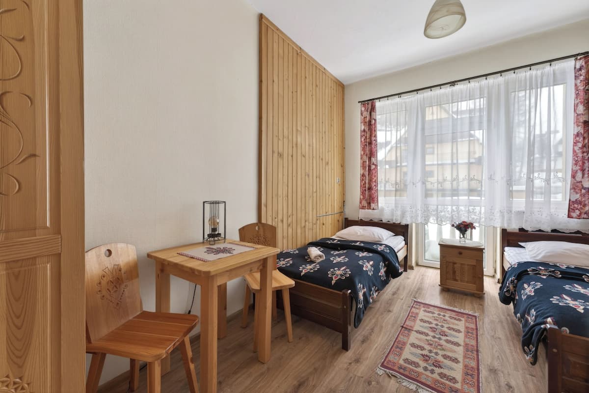 A bedroom with two beds, a wooden desk, a chair, a nightstand, and a window with curtains.