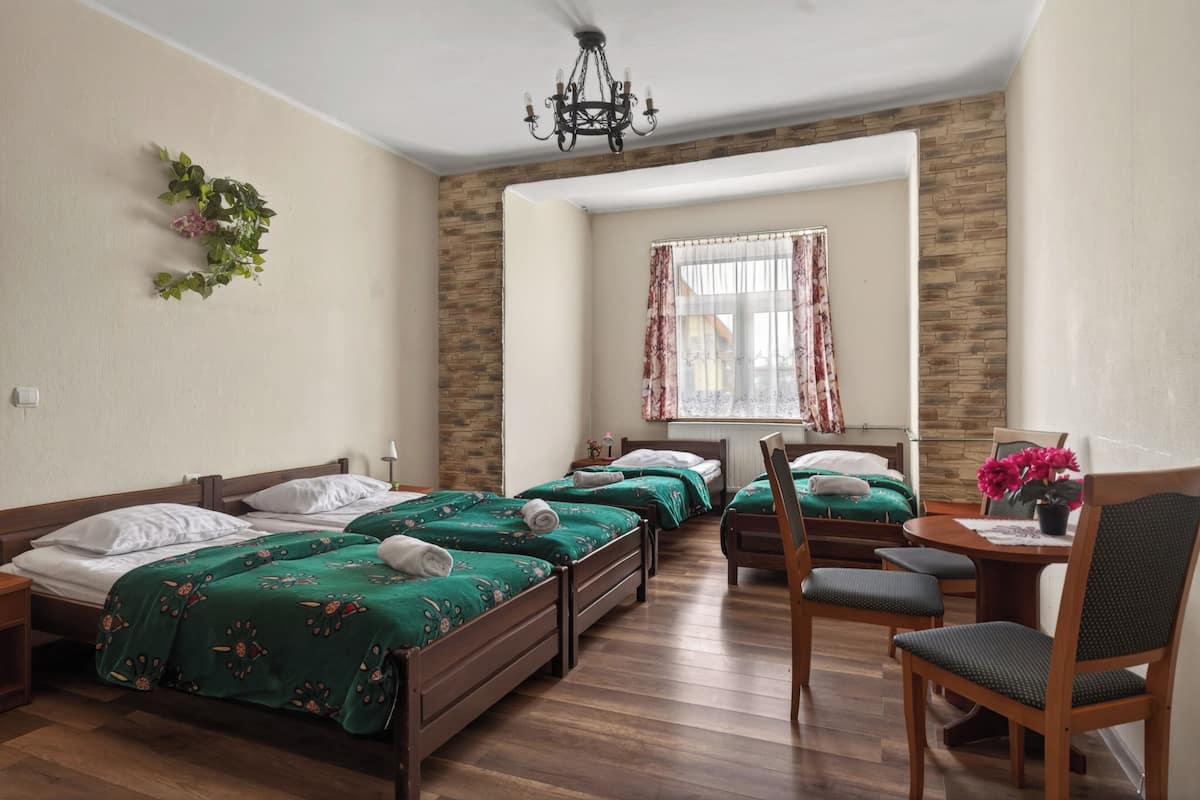 A hotel room with four single beds, a wooden floor, a chandelier, a window with curtains, and a small table with a chair.