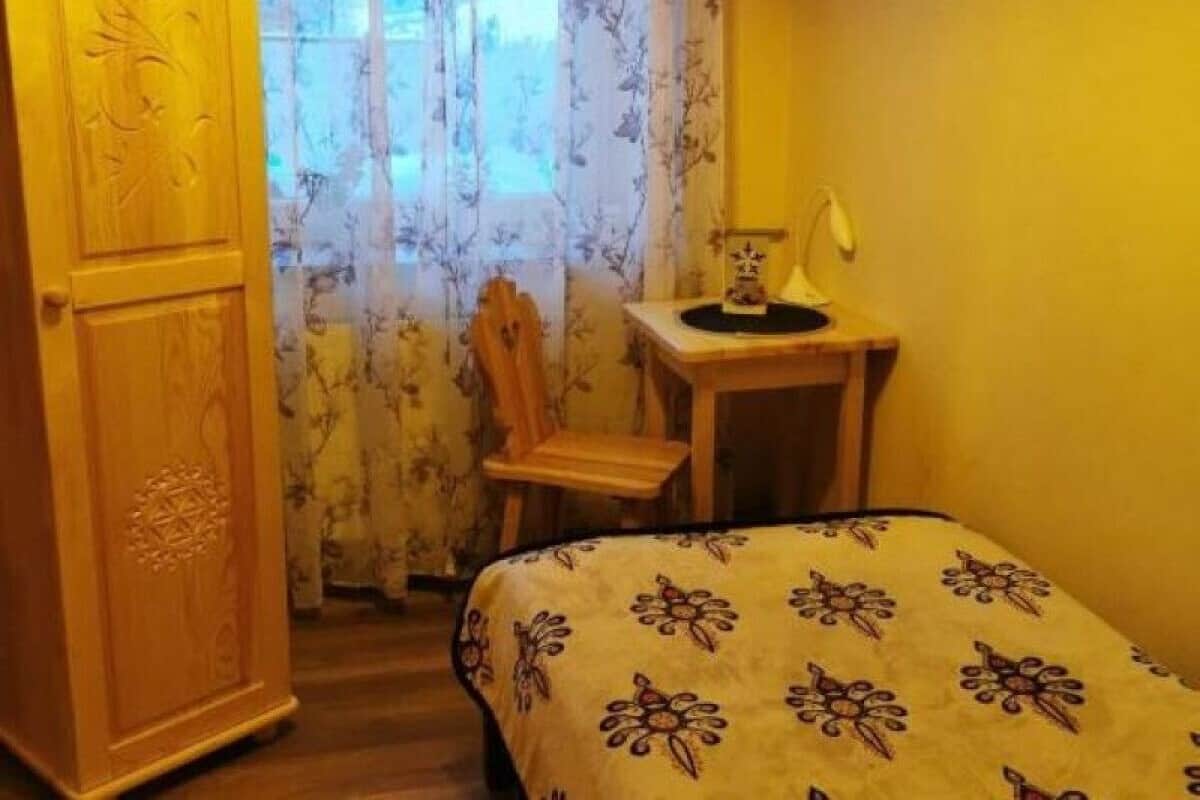 A bedroom with a bed, a wooden wardrobe, a desk with a lamp, and a chair.