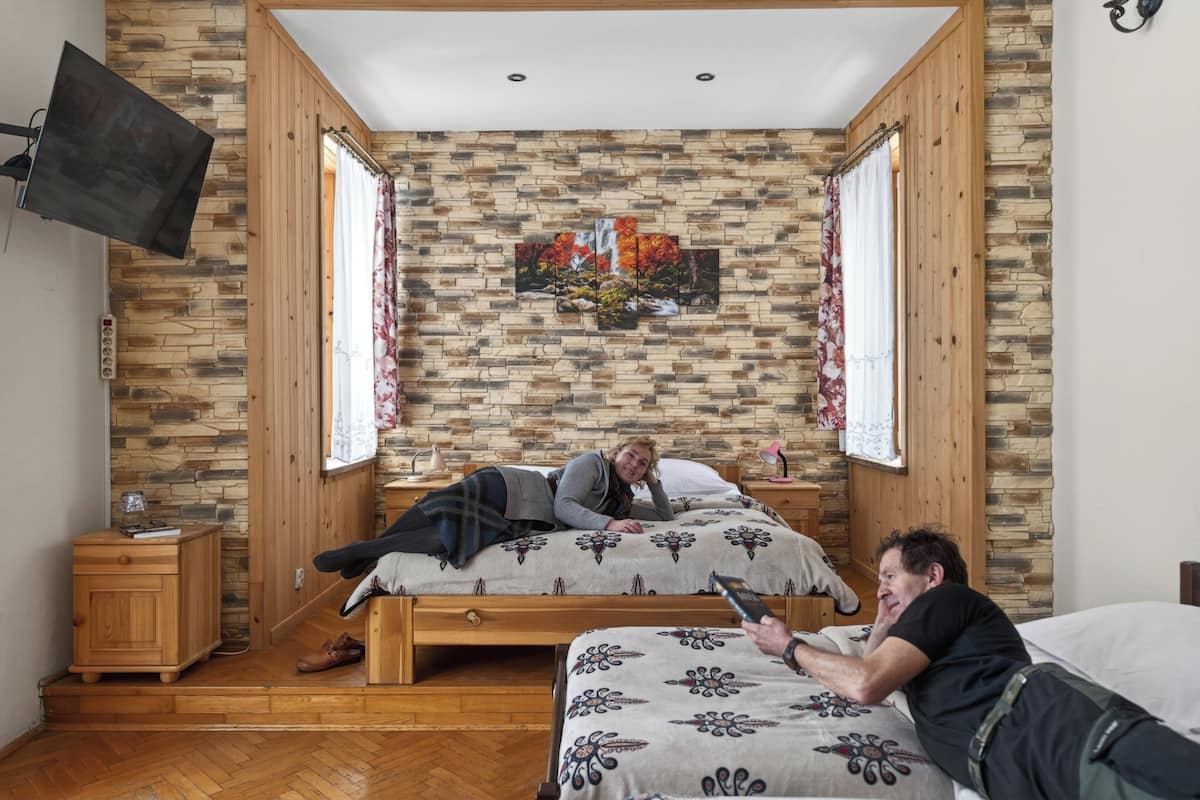 A bedroom with a stone wall, wooden paneling, a bed with a patterned bedspread, a bedside table, and a wall-mounted TV.