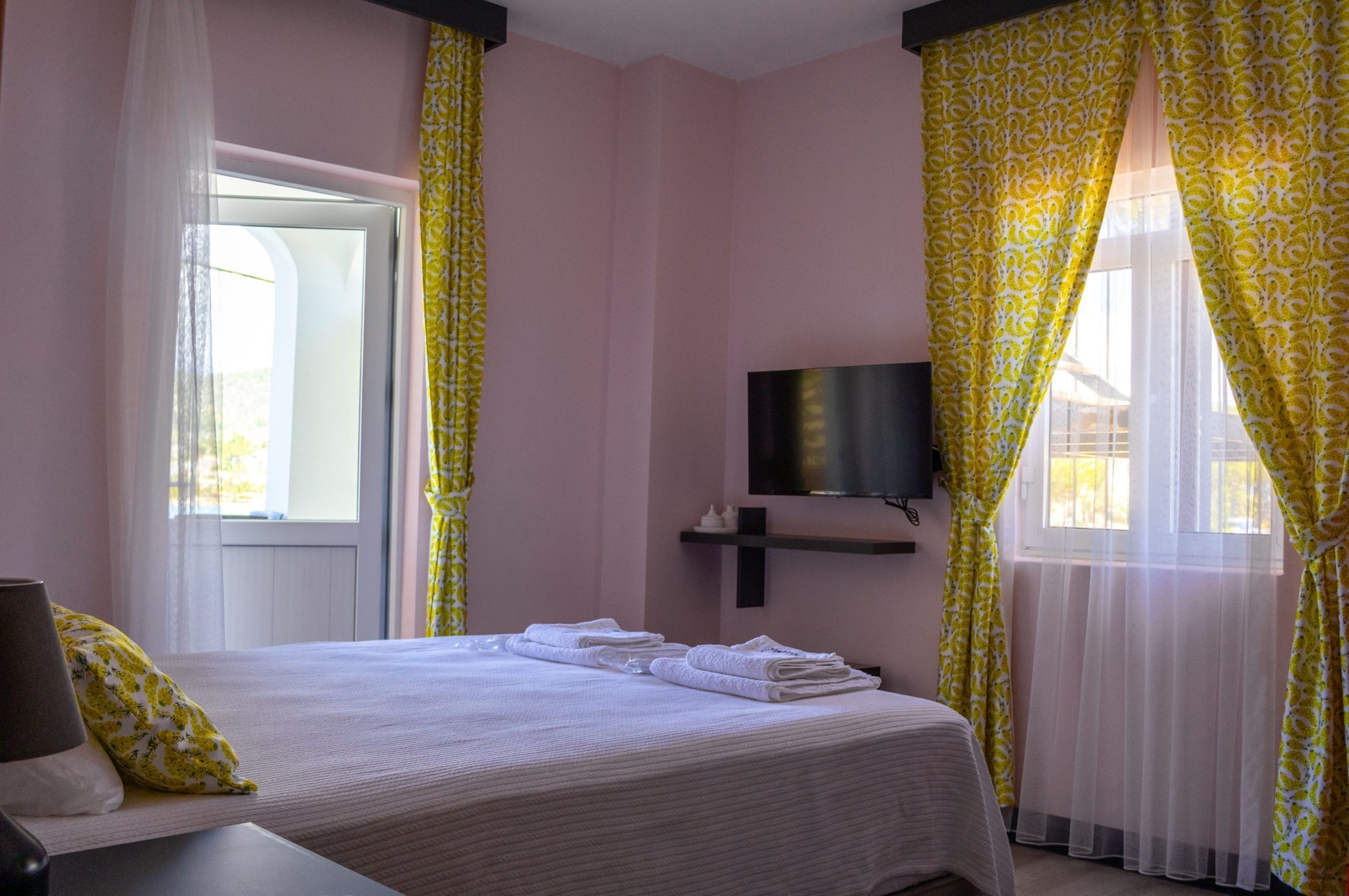 A hotel room with a bed, a TV mounted on the wall, and a window with yellow curtains.