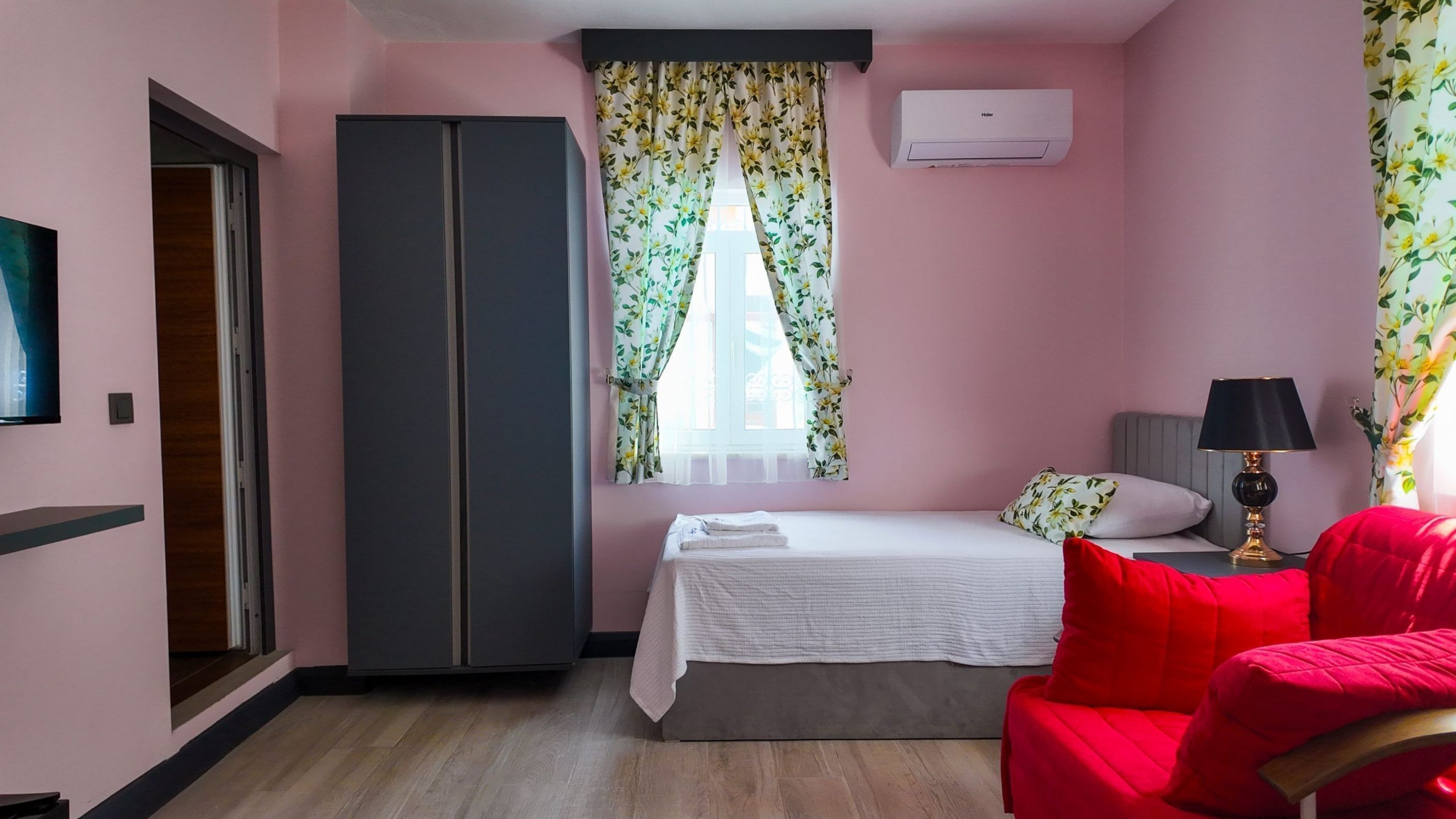 A bedroom with a bed, a red chair, a wardrobe, a window with floral curtains, and an air conditioning unit.