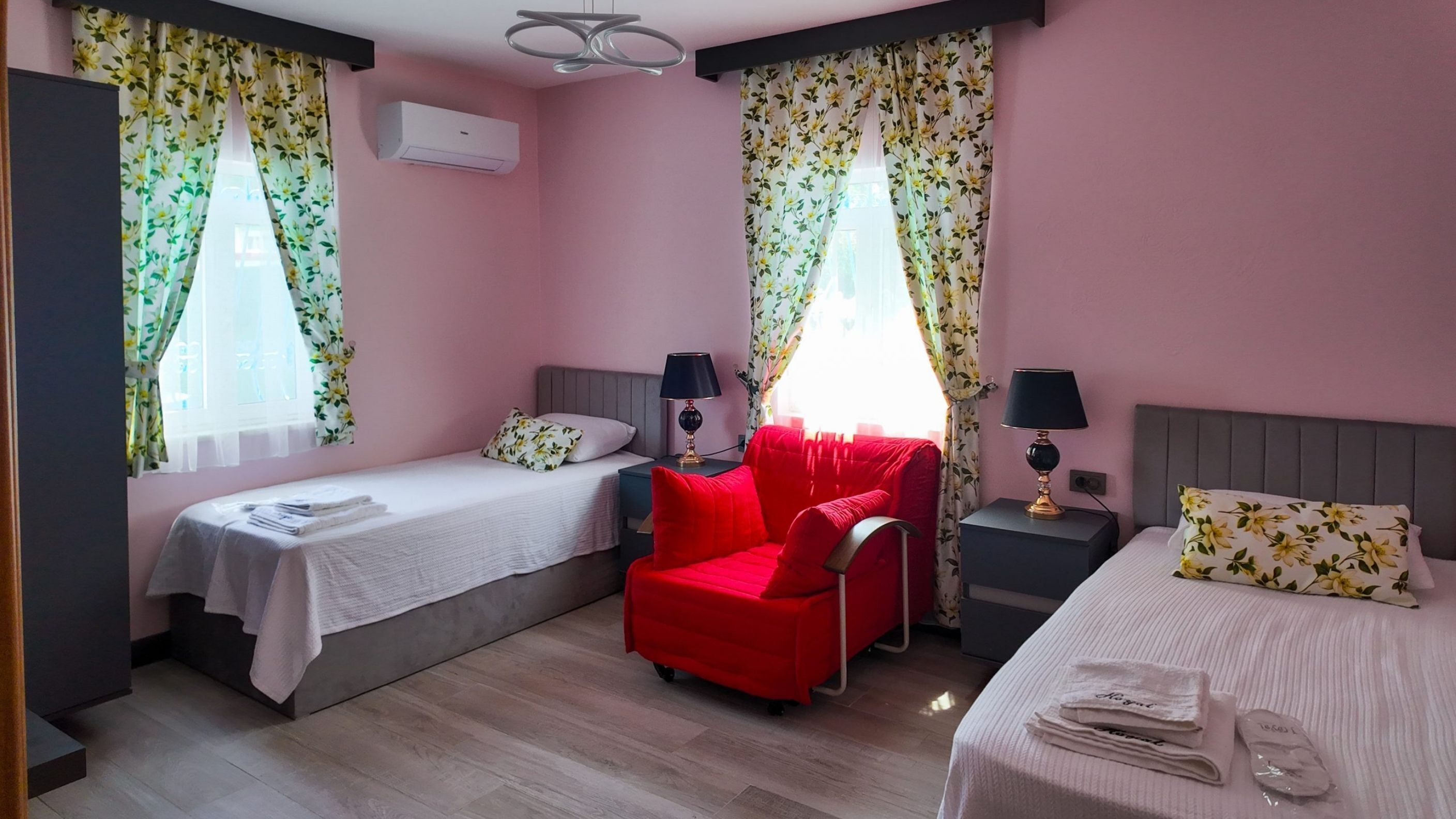 Habitación de hotel con dos camas individuales, un sillón rojo, un escritorio con lámpara y una ventana con cortinas florales.