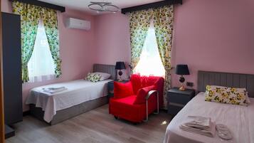 Habitación de hotel con dos camas individuales, un sillón rojo, un escritorio con lámpara y una ventana con cortinas florales.