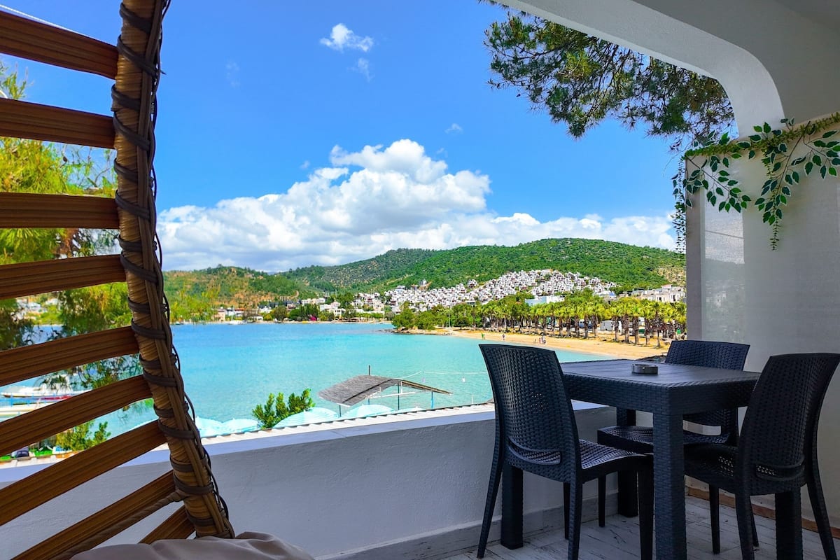 A balcony with wicker furniture overlooking a beach and a hill with white buildings.