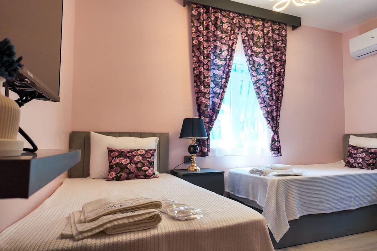 A bedroom with two beds, a nightstand with a lamp, a window with floral curtains, and an air conditioner on the wall.