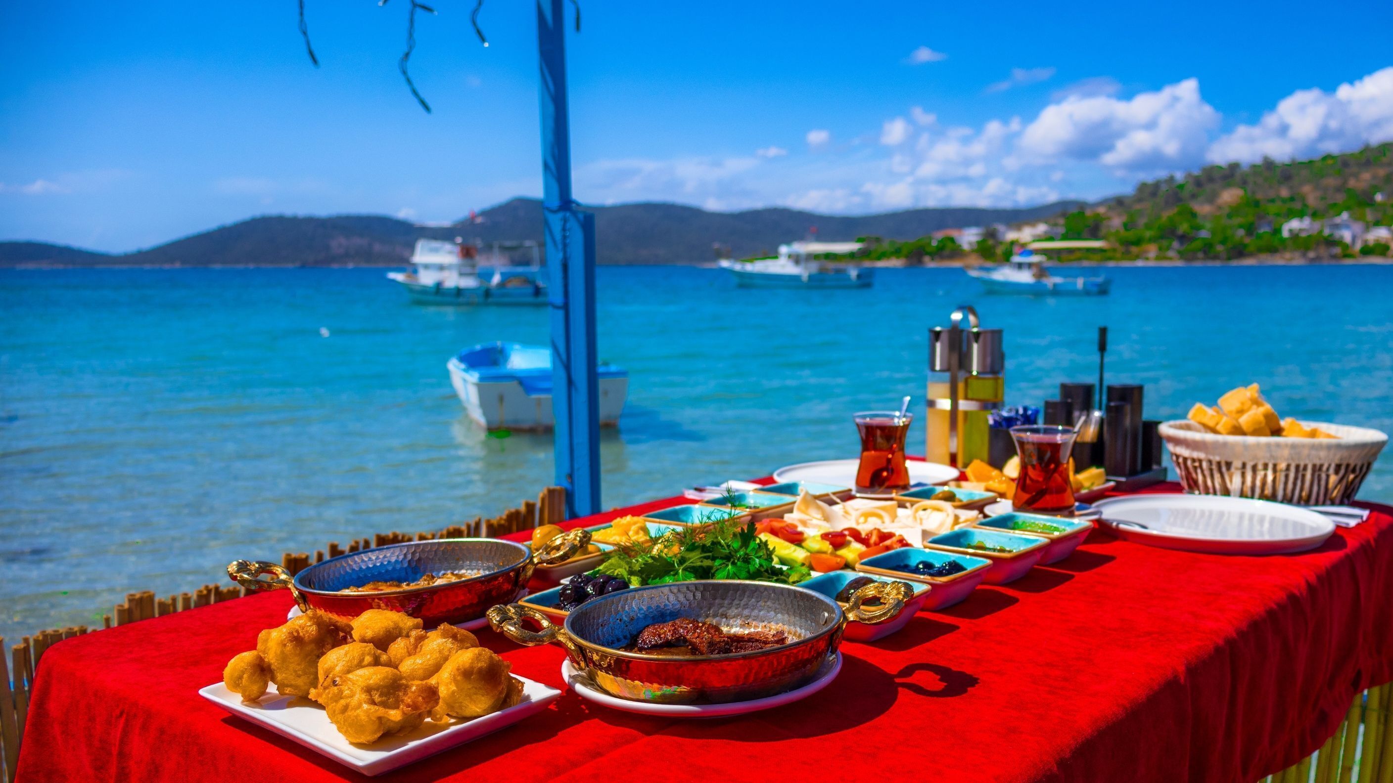 A table set with various dishes and drinks, overlooking a body of water with boats and a mountainous landscape in the background.