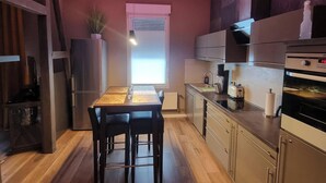 Eine moderne Küche mit Holzboden, einem Esstisch mit Stühlen, einem Kühlschrank, einem Herd und einem Fenster mit Jalousien.