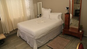 A neatly made bed with white linens, a wooden dresser with a mirror, and a small bedside table with a lamp. - Coco Palm B&B (Windhoek)